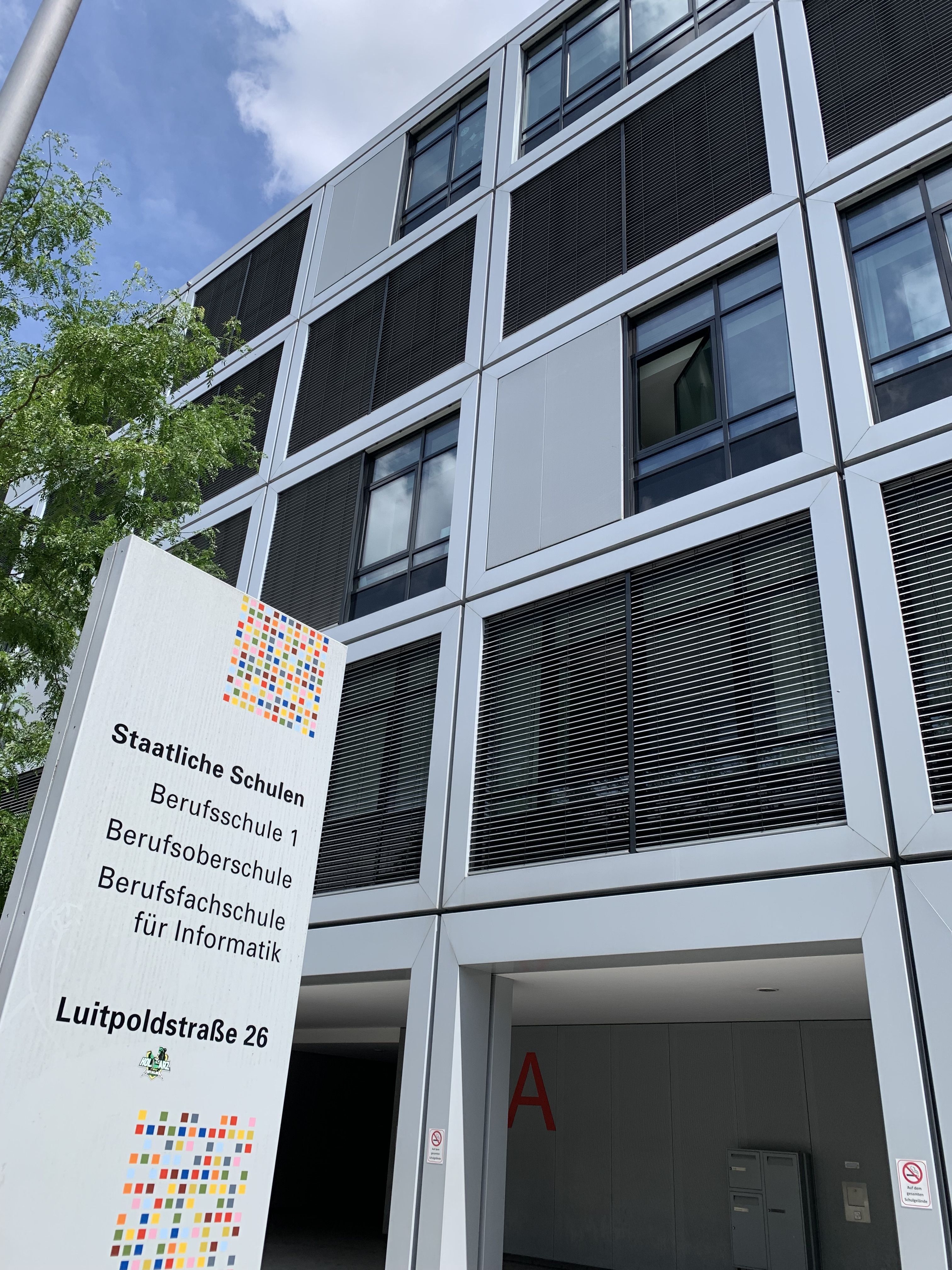 Es ist ein Gebäudeteil der Berufsschule 1, Luitpoldstraße 26 in 84034 Landshut zu sehen. Im linken Vordergrund ist eine Stele mit den Schulbezeichnungen sowie der Adresse.